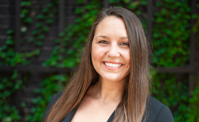 Kim Beechner, owner of Embark Marketing, a marketing agency based in Lubbock, Texas. She is smiling, has long straight brown hair, and is wearing a black top and delicate necklace. The background features a dark wooden trellis with green ivy.