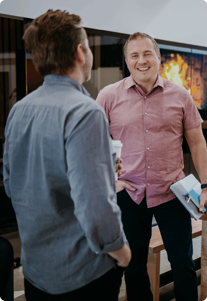 Russel Dubree, smiling in conversation with another person while holding a notebook, wearing a pink button-down shirt in an office setting.