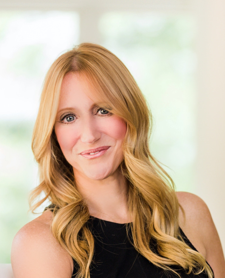 Lori Asbury, owner of CMOco, sits comfortably on a white chair in a softly lit space with a blurred window in the background. She wears a sleeveless black top, white pants, and neutral-toned heels. Her long, blonde hair is styled in soft waves, and she accessorizes with a silver bracelet and ring. Lori smiles warmly with a confident, composed presence.