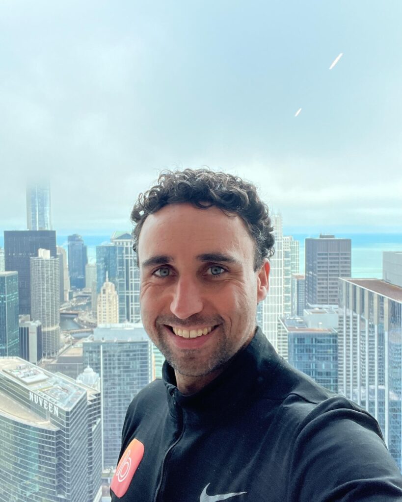 Luke Perrie, owner of Bandicoot Marketing, takes a selfie in front of a high-rise window with a stunning view of downtown Chicago’s skyline. He wears a black Nike jacket featuring the Bandicoot logo and smiles brightly at the camera. Tall buildings and a misty sky over Lake Michigan form the dramatic cityscape backdrop.