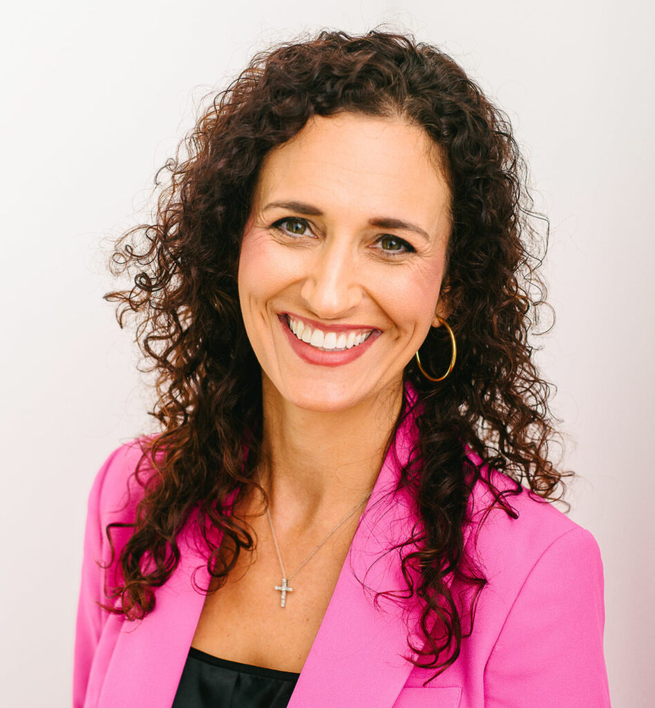 Rachel Ledet, owner of 30|90 Marketing, smiles warmly in front of a clean white background. She wears a vibrant pink blazer over a black top, paired with gold hoop earrings and a silver cross necklace. Her curly dark hair frames her face, and her confident, approachable demeanor shines through in this professional portrait.