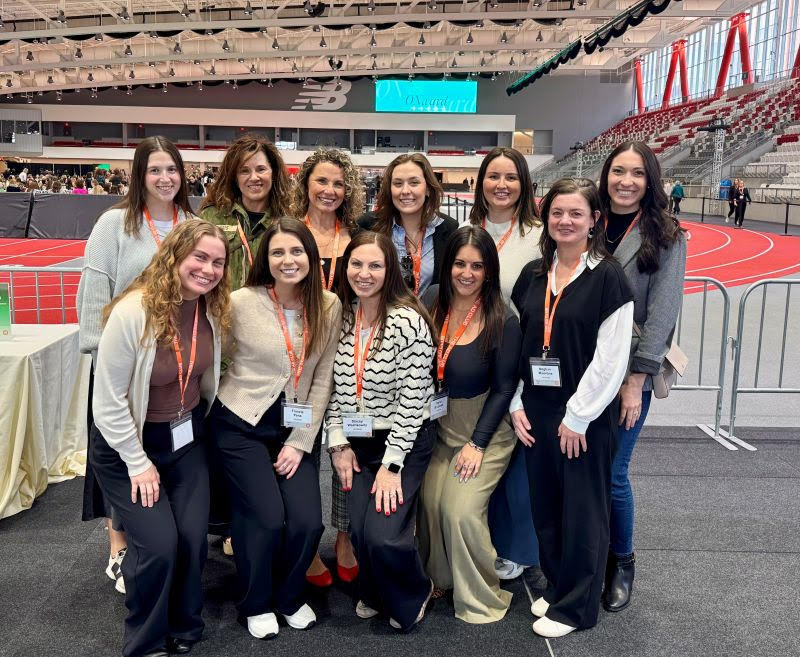 Group photo of eleven women wearing conference badges and orange lanyards, smiling together inside a large indoor sports and event venue with a running track in the background.